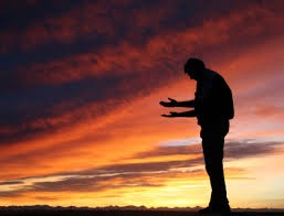 A man bowing his head with his arms raised to heaven. The sky is filled with the colors of a sunset.